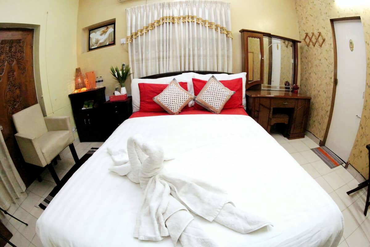 A neatly made bed with a white comforter, red pillows, and a folded white towel. There is a wooden dresser with a mirror, a chair, and a small table with a plant.