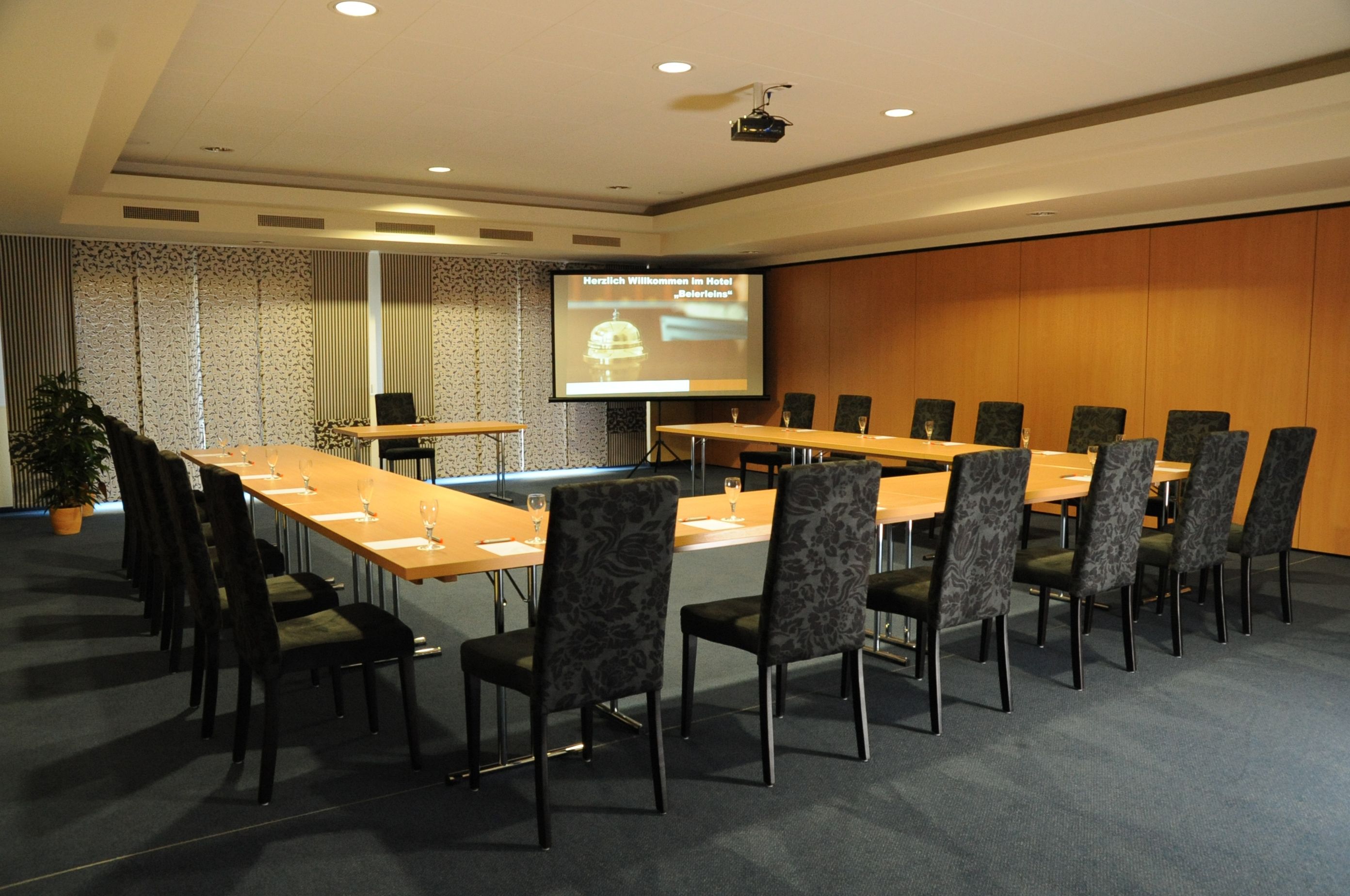 A conference room with a long table, chairs, a projector screen, and a potted plant.