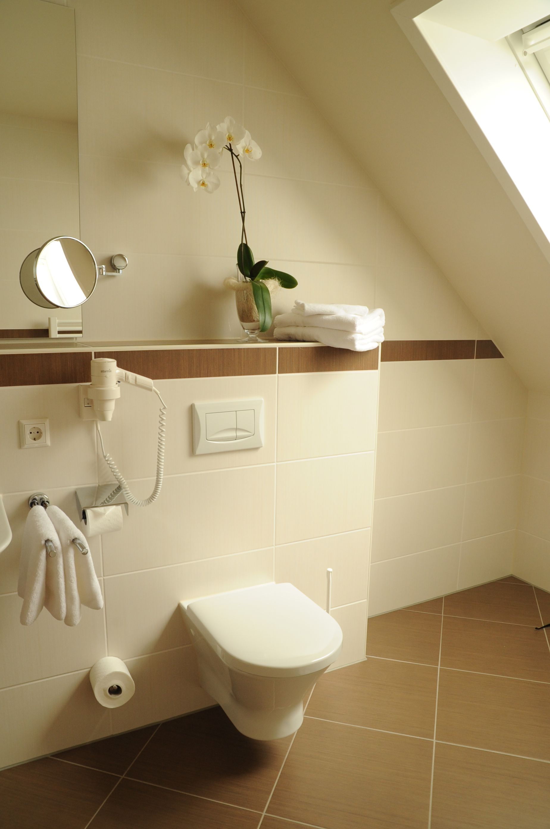 Une salle de bain équipée de toilettes, d’un porte-serviettes, d’un sèche-cheveux, d’un miroir et d’une orchidée en pot.