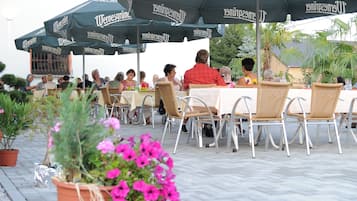 An outdoor dining area with tables and chairs under umbrellas, surrounded by potted plants and a pool.