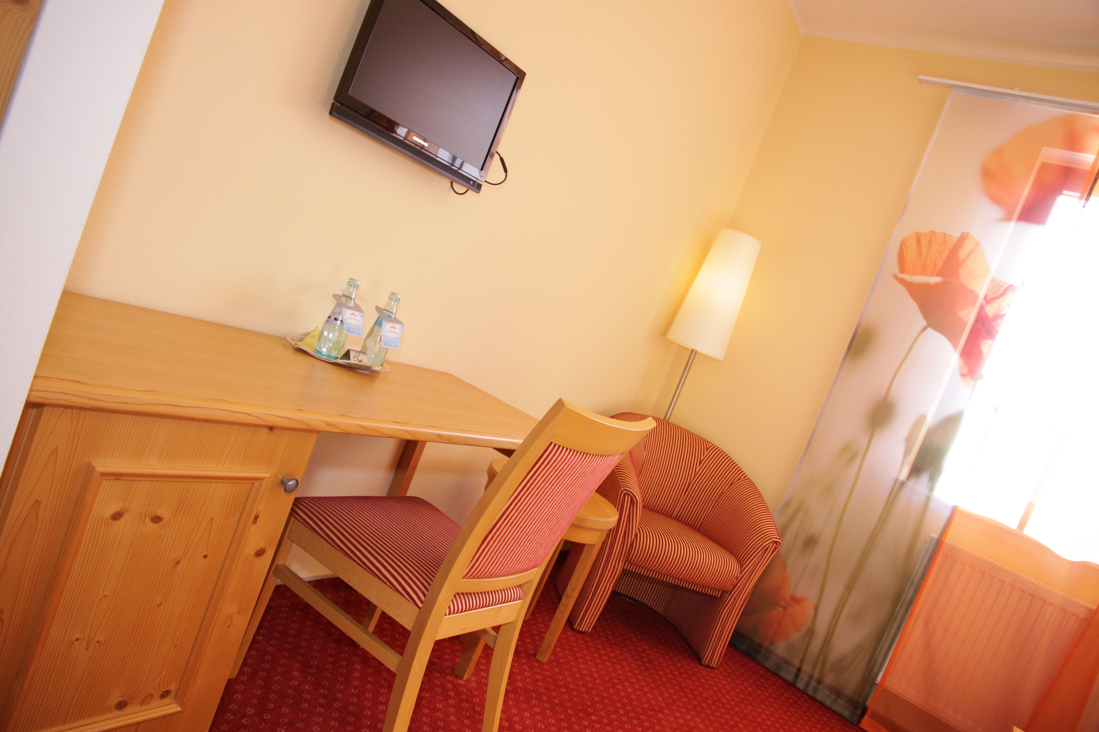 Habitación de hotel con un escritorio de madera, dos sillas, un televisor montado en la pared, una lámpara y una ventana con cortina estampada.