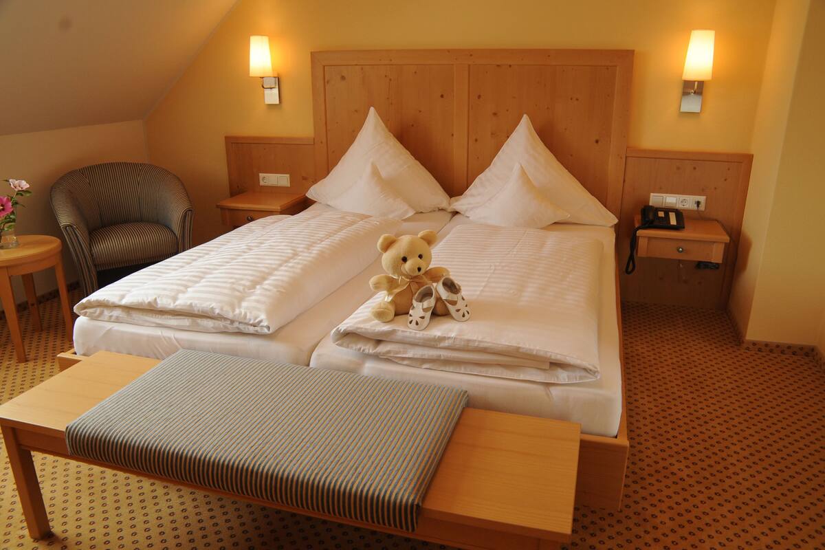 A double bed with pillows, a bedside table, a chair, a bedside lamp, and a teddy bear on the bed.