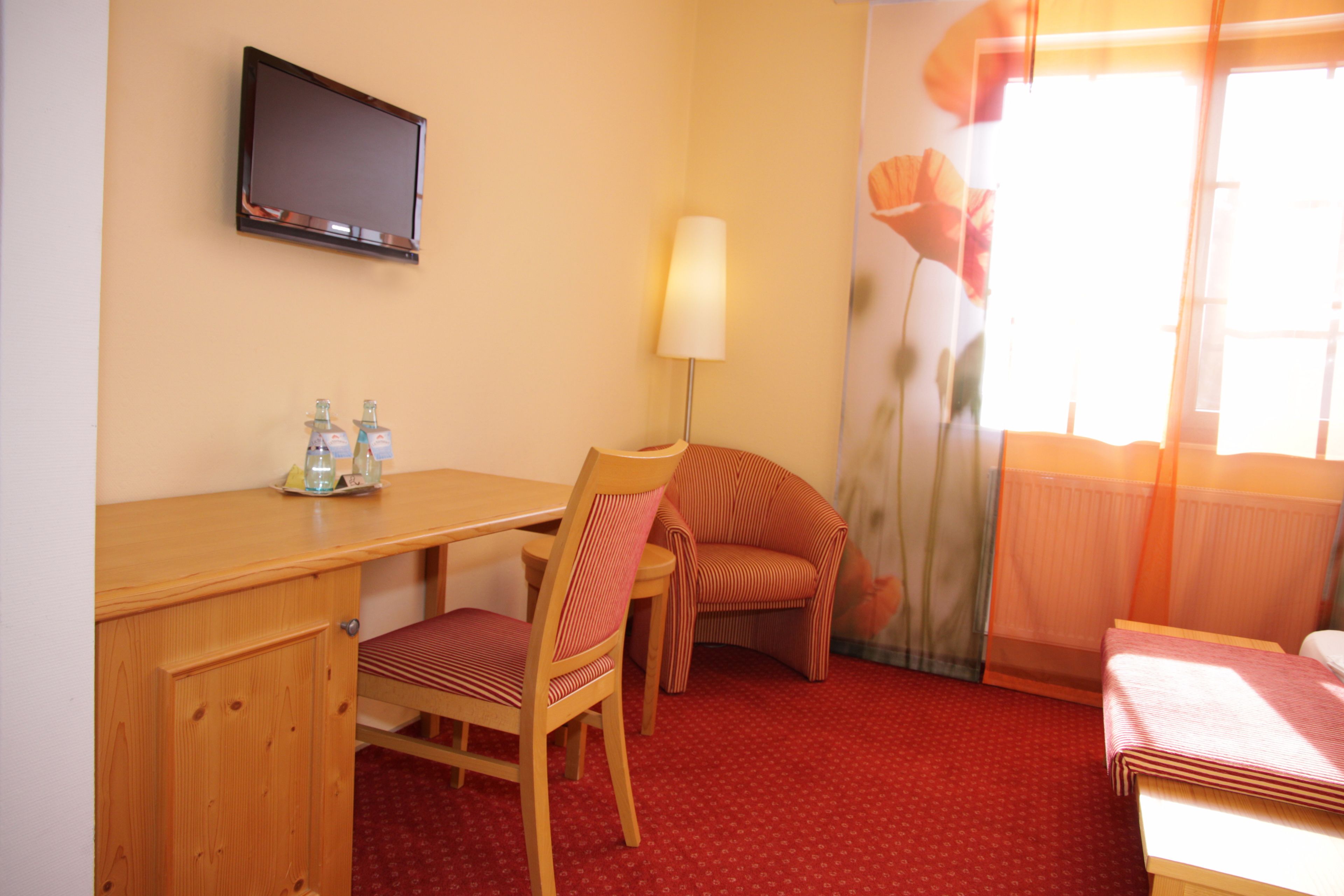 A hotel room with a wooden desk, a chair, a bed, a television, and a window with curtains.