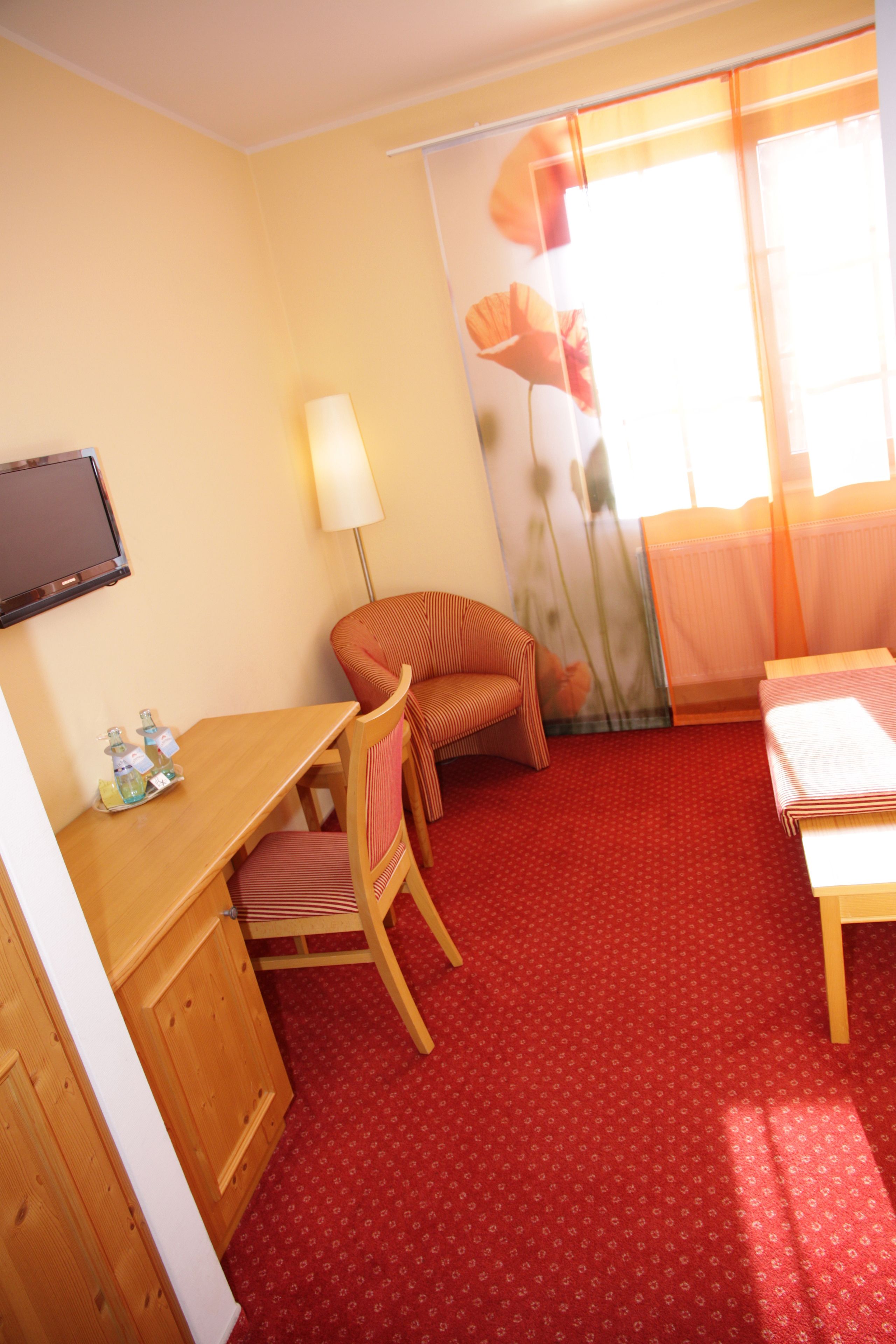 Ein Hotelzimmer mit einem Holztisch, einem Sessel, einem Bett, einem an der Wand befestigten Fernseher und einem Fenster mit Vorhängen.