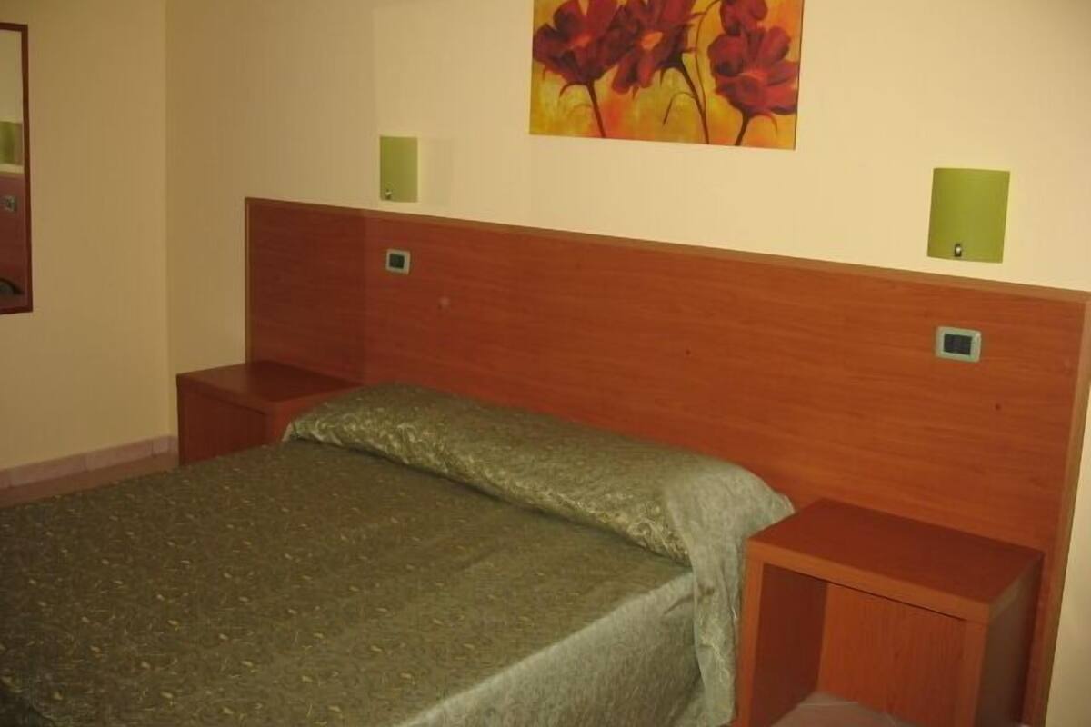 A hotel room with a wooden headboard, a single bed with a green bedspread, two bedside tables with green lamps, and a painting of red flowers on the wall.