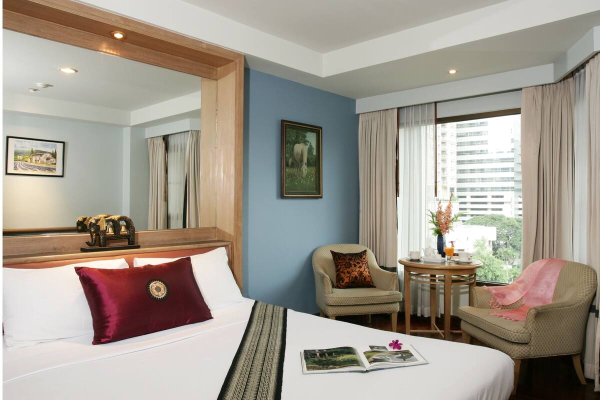 Habitación de hotel con una cama grande, una silla, una mesita con un florero y una ventana con cortinas.