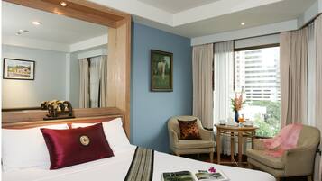 Quarto de hotel com uma cama grande, uma cadeira, uma mesinha com um vaso e uma janela com cortinas.