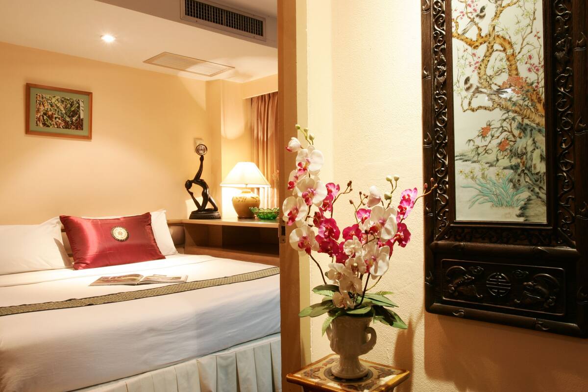 Habitación de hotel con cama, mesita de noche con lámpara, un jarrón con flores y un cuadro en la pared.
