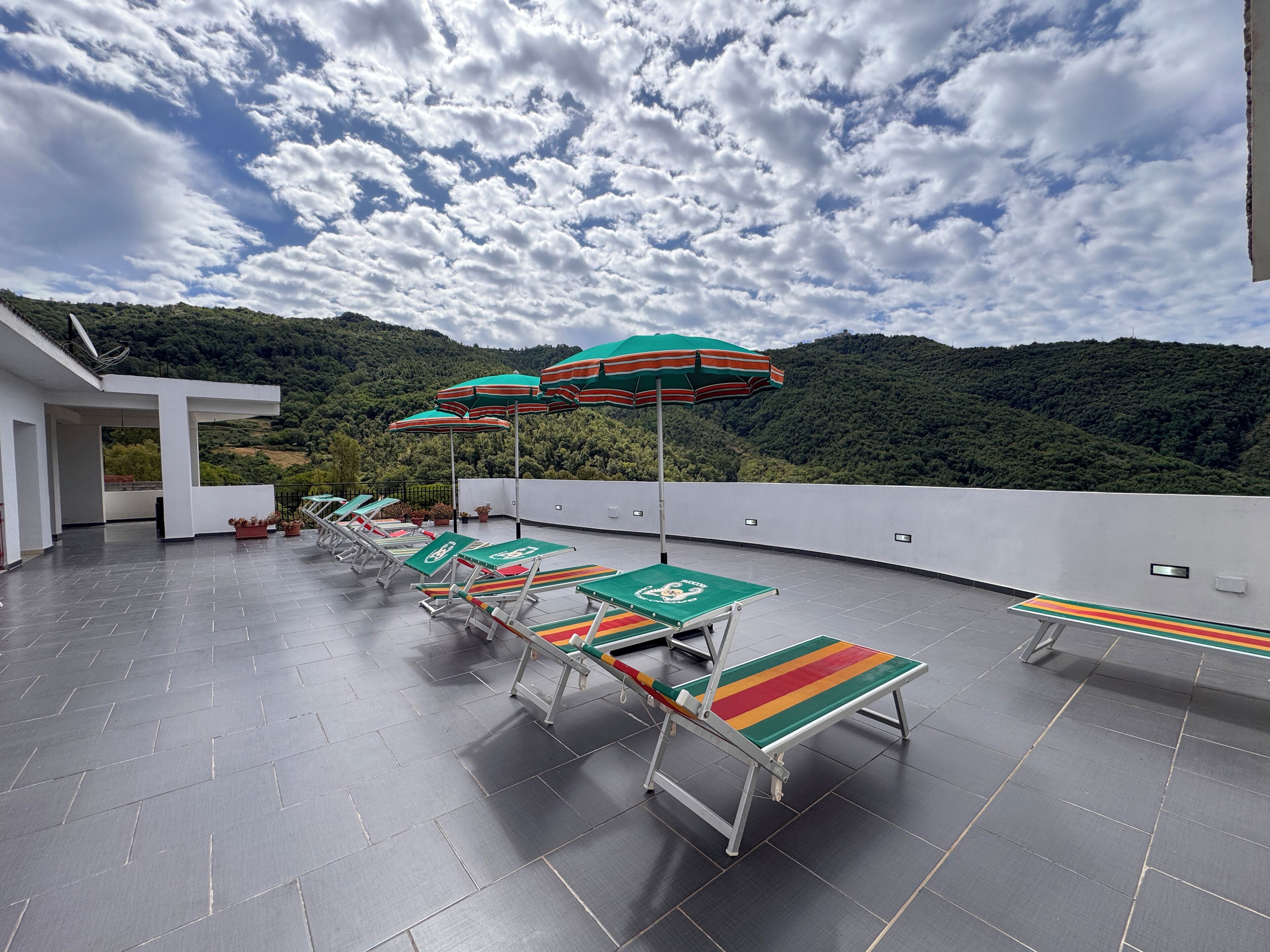 Une terrasse sur le toit, dotée de plusieurs chaises longues et de parasols, un bâtiment blanc et un paysage montagneux en arrière-plan.