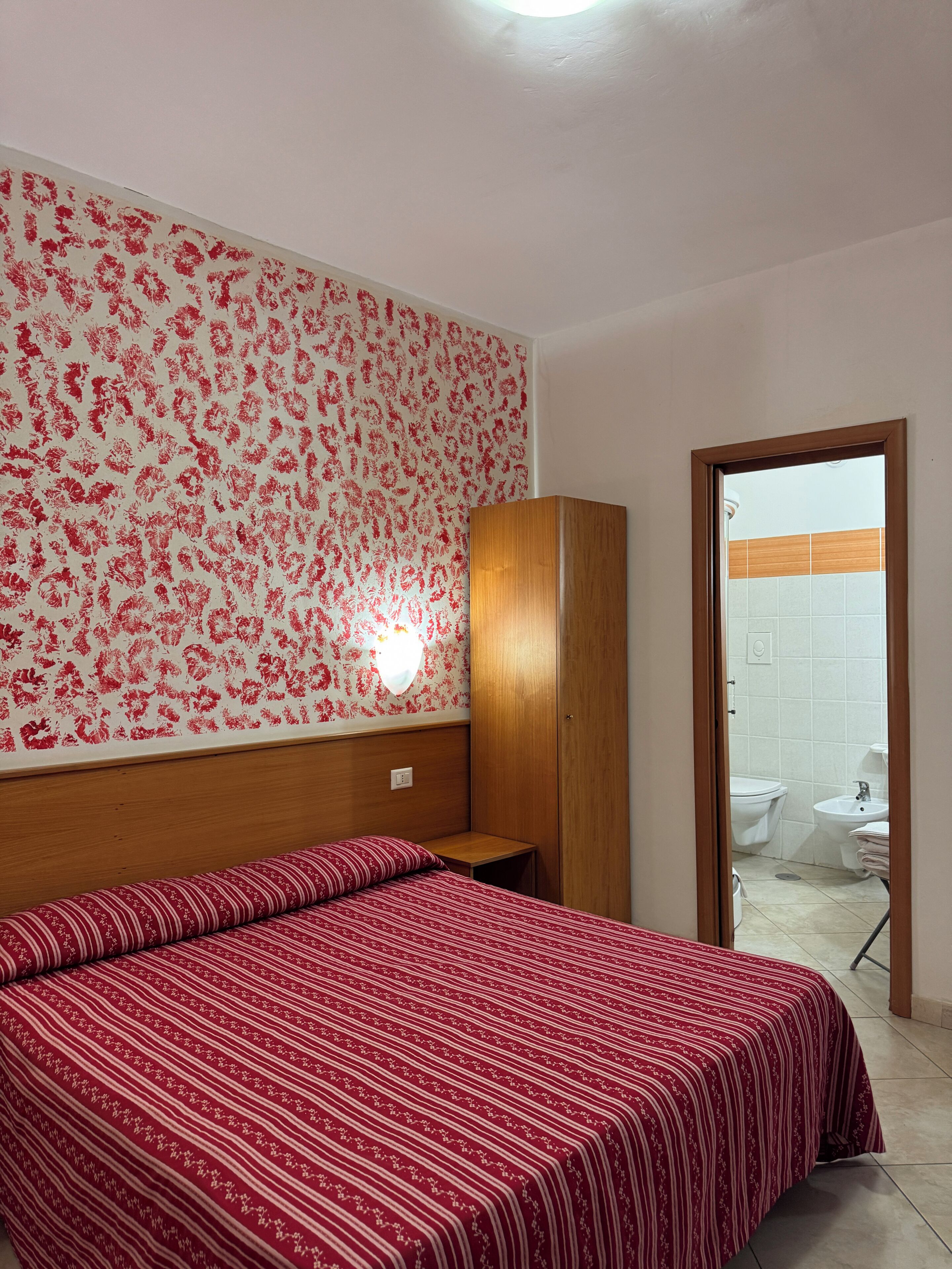 Ein Schlafzimmer mit Bett, Kleiderschrank, Wand mit Blumenmuster und angrenzendem Badezimmer, das durch eine offene Tür sichtbar ist.