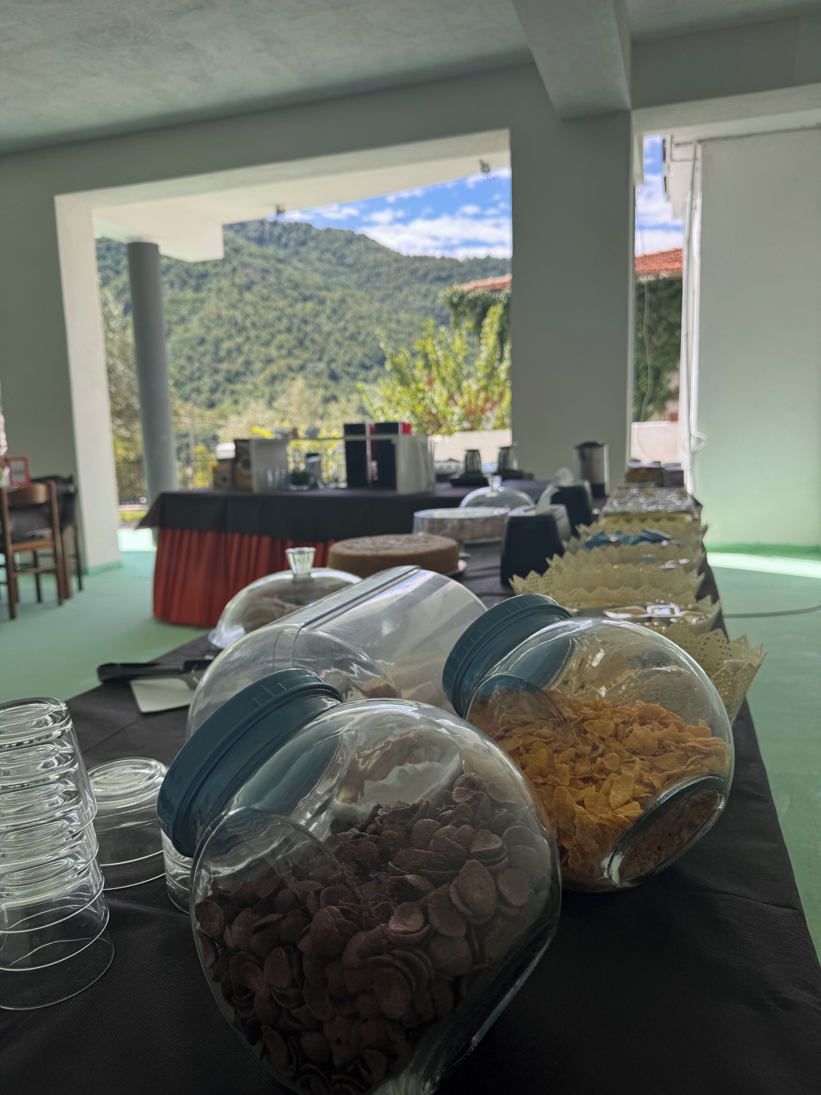 Una mesa de buffet con diversos recipientes de comida y vista a un paisaje montañoso a través de una puerta abierta.