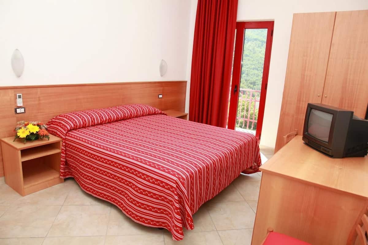 Un dormitorio con una cama, un televisor, un armario de madera, una mesita de noche con un florero y una cortina roja.