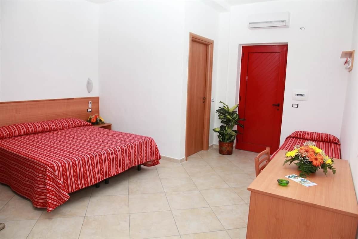 Habitación de hotel con cama, escritorio, silla, puerta roja y planta en maceta.