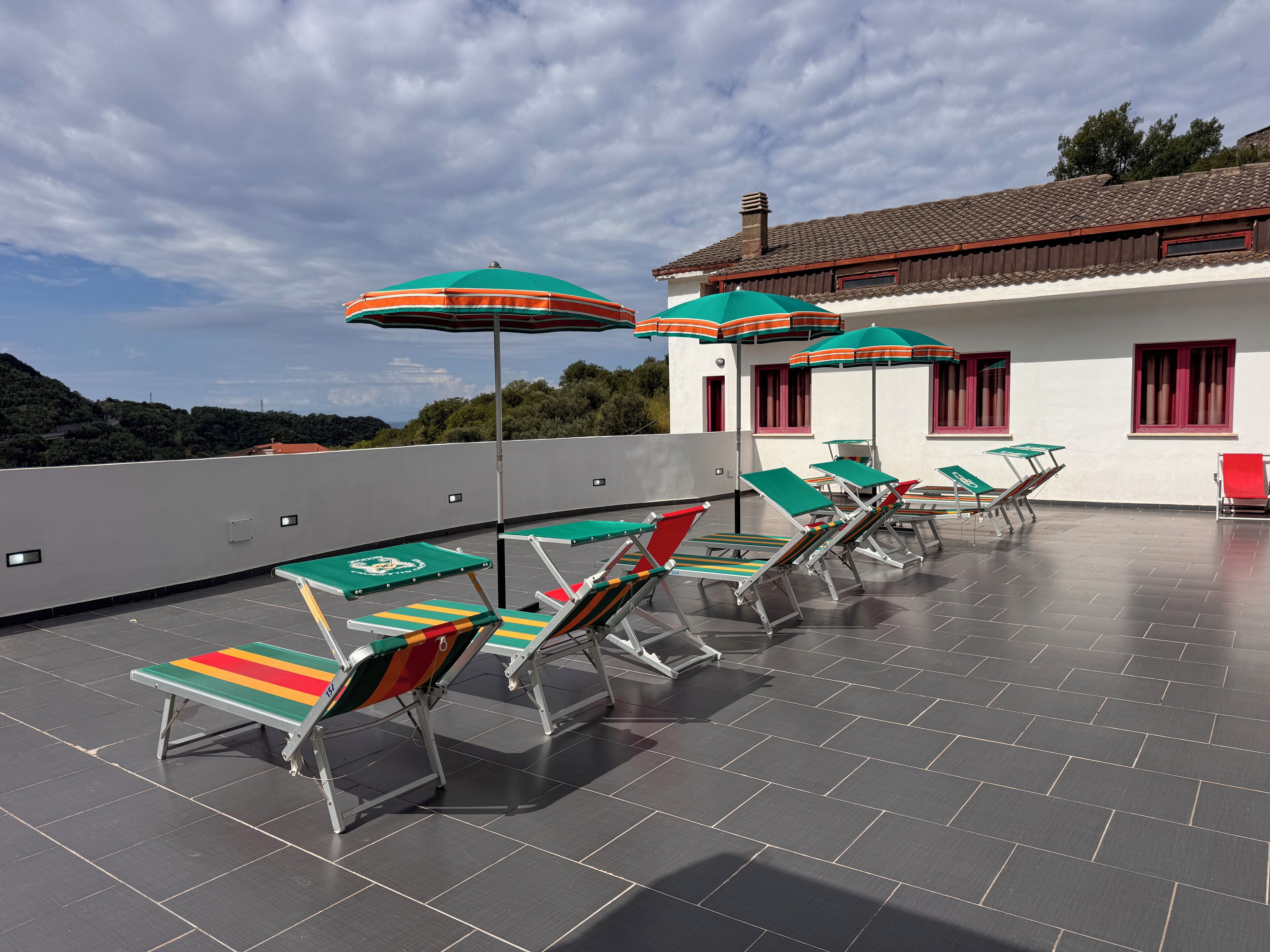 Eine Dachterrasse mit Liegestühlen und Sonnenschirmen, ein weißes Gebäude mit roten Fensterrahmen und Blick auf die Landschaft.