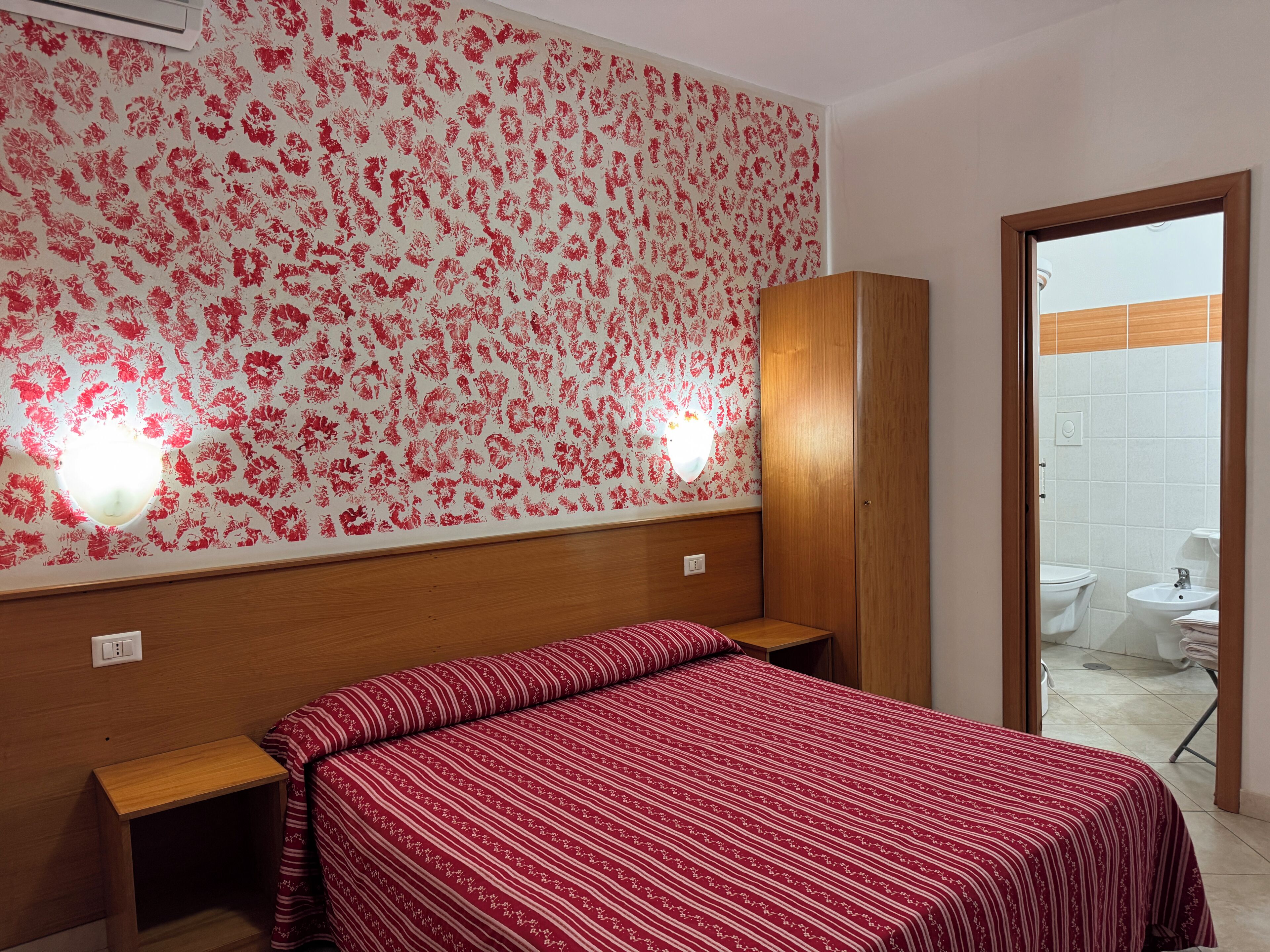 A hotel room with a bed, a wooden headboard, a wall with a floral pattern, and a bathroom visible through an open door.