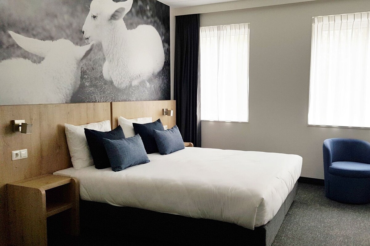 A hotel room with a large bed, a chair, a small table, and a painting on the wall.