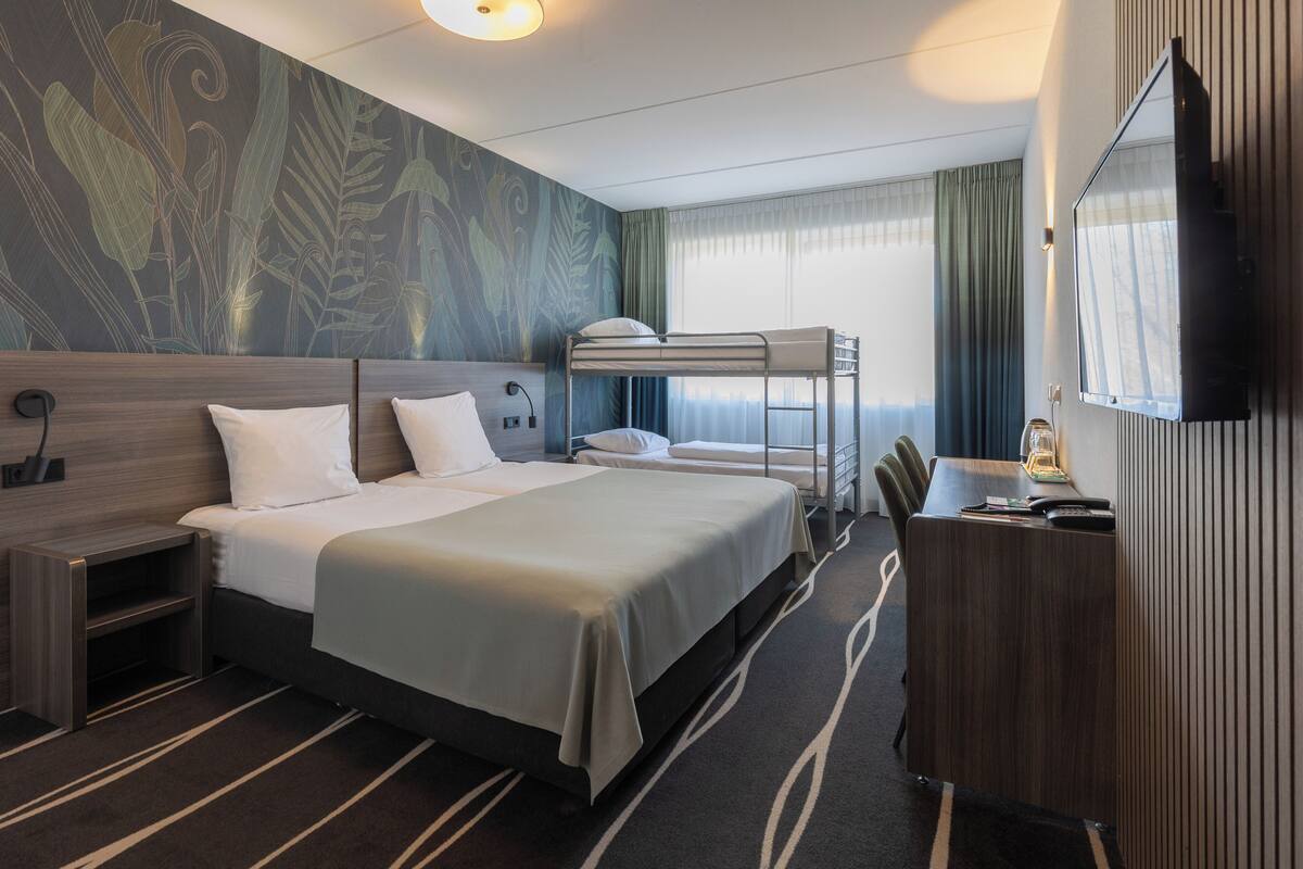 Une chambre d’hôtel avec un lit superposé, un bureau avec une lampe, une télévision et une grande fenêtre avec des rideaux.