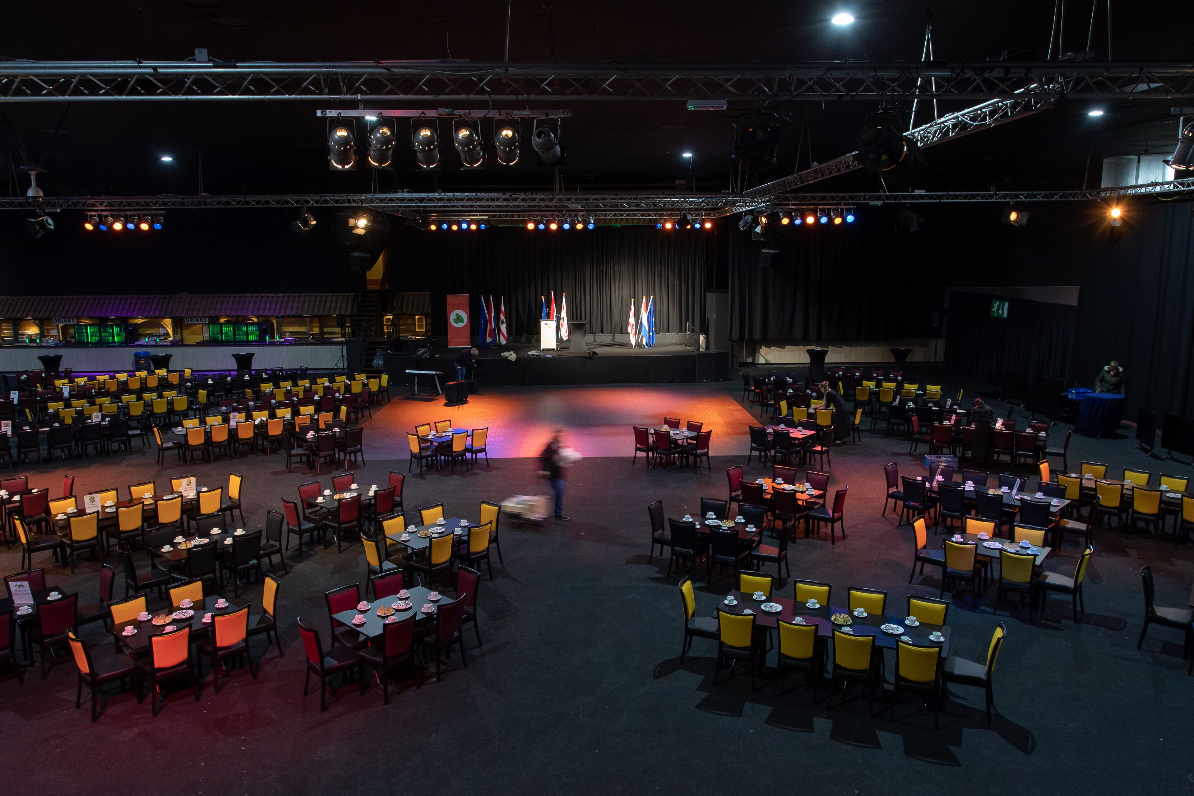 A large indoor venue with multiple dining tables arranged in rows, a stage with a curtain backdrop, and flags on either side.