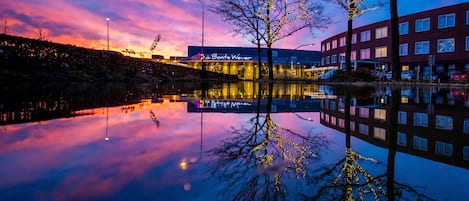 A building with a sign that reads "De Bontje Wever" reflecting in a calm body of water, surrounded by trees and a clear sky.