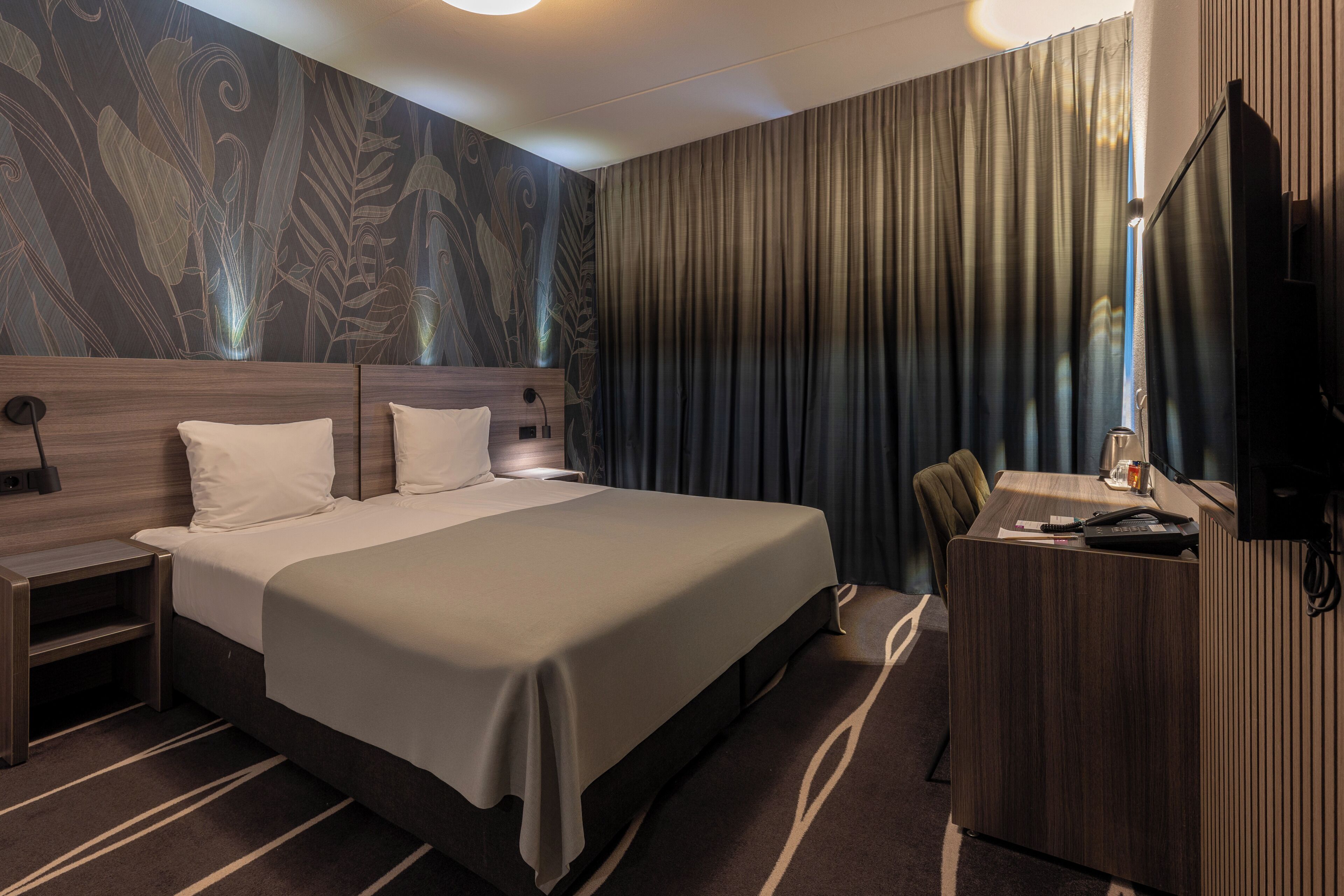 Hotelkamer met een groot bed, een bureau, een stoel, een televisie en een wand met een bladpatroon.