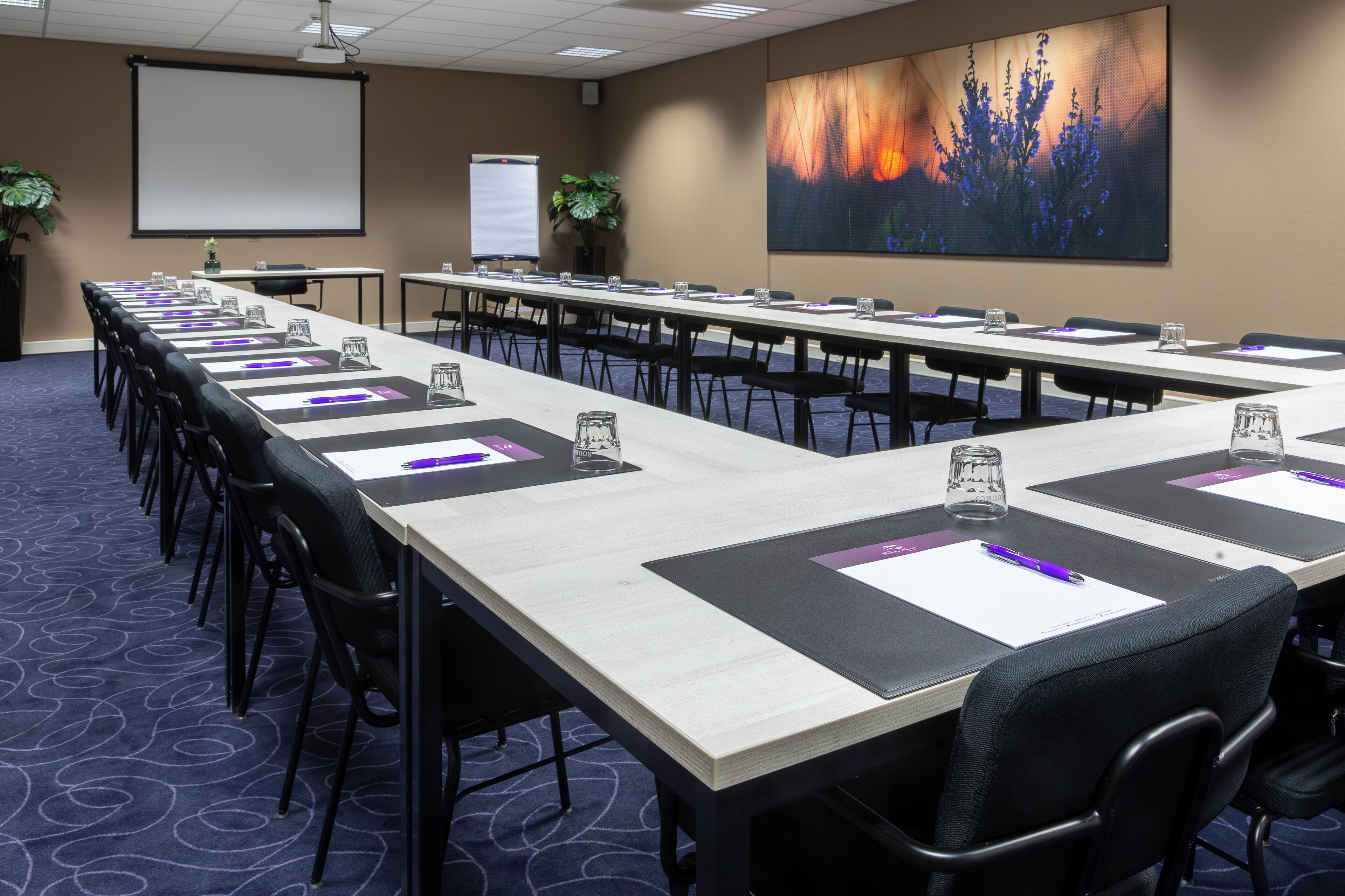 Une salle de conférence avec de longues tables, des chaises, un écran de projection et un grand tableau abstrait au mur.