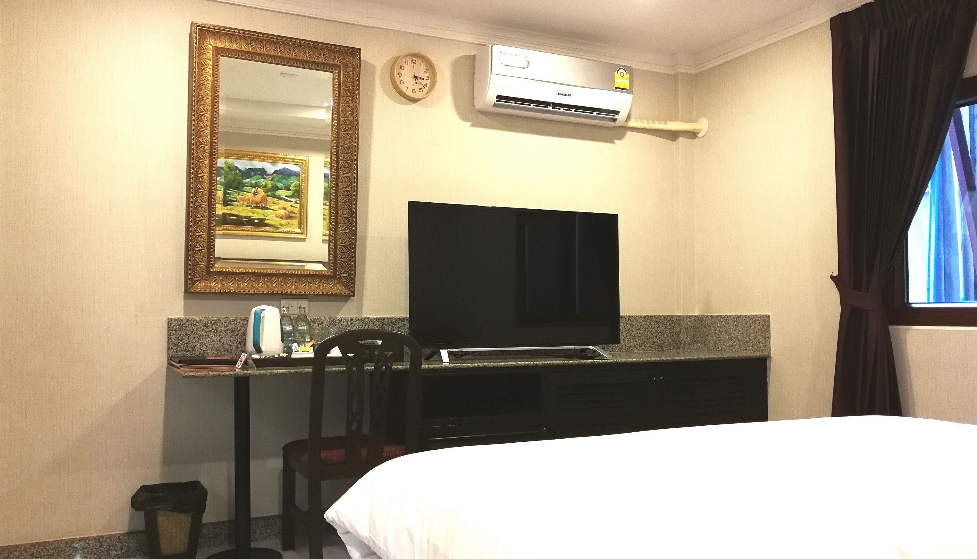 A hotel room with a bed, a flat-screen TV, a small table with a kettle, a chair, a clock, and an air conditioning unit.