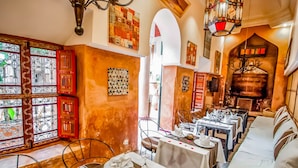Un restaurant avec des tables dressées pour le dîner, un lustre et une vue sur l’extérieur à travers une fenêtre ornée de ferronnerie décorative.