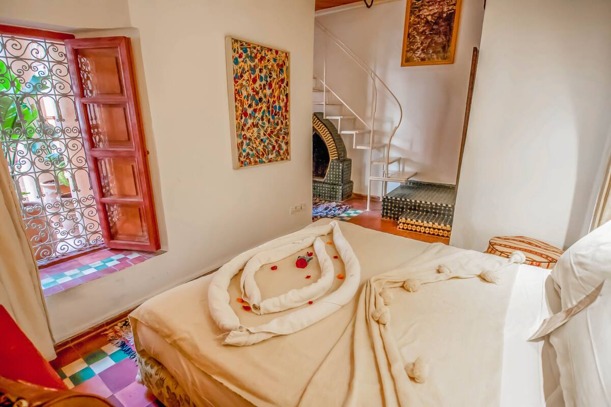Een slaapkamer met een bed, een raam met decoratieve ijzeren spijlen, een trap en een muur met een kleurrijk abstract schilderij.