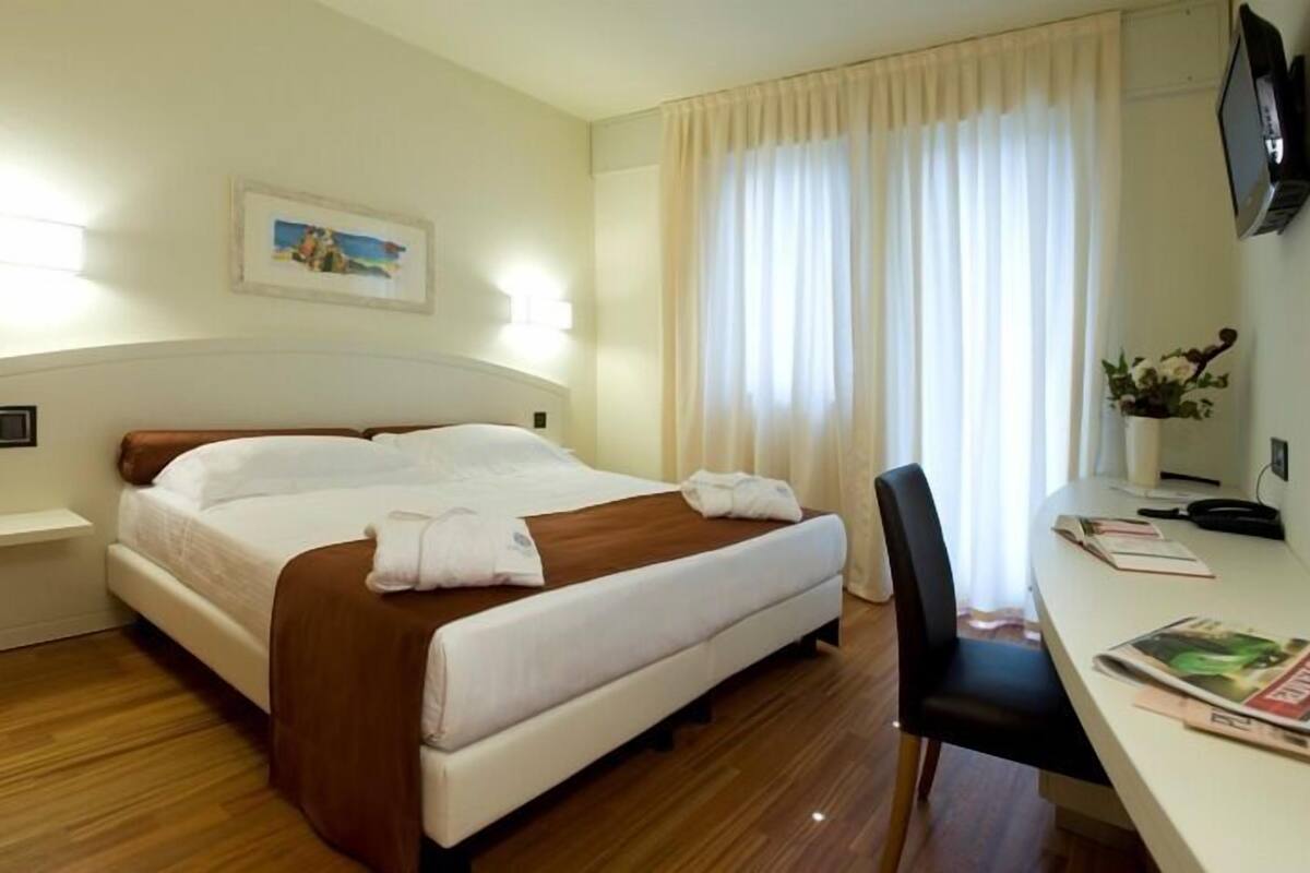 A hotel room with a bed, bedside table, chair, desk, and a TV on the wall.