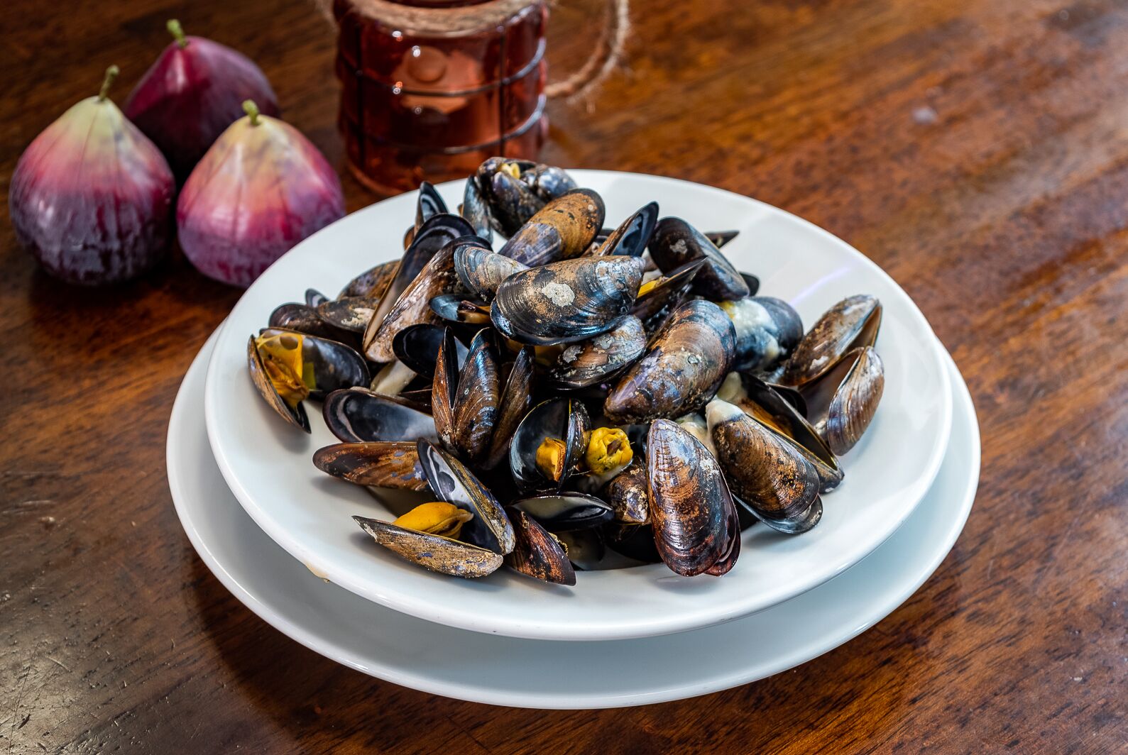 Un plato de mejillones sobre un plato blanco, con dos bulbos de ajo enteros y una taza de cobre al fondo.