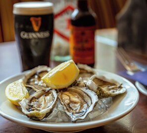 A plate of oysters with lemon wedges on a table, accompanied by a glass of beer and a bottle of sauce.