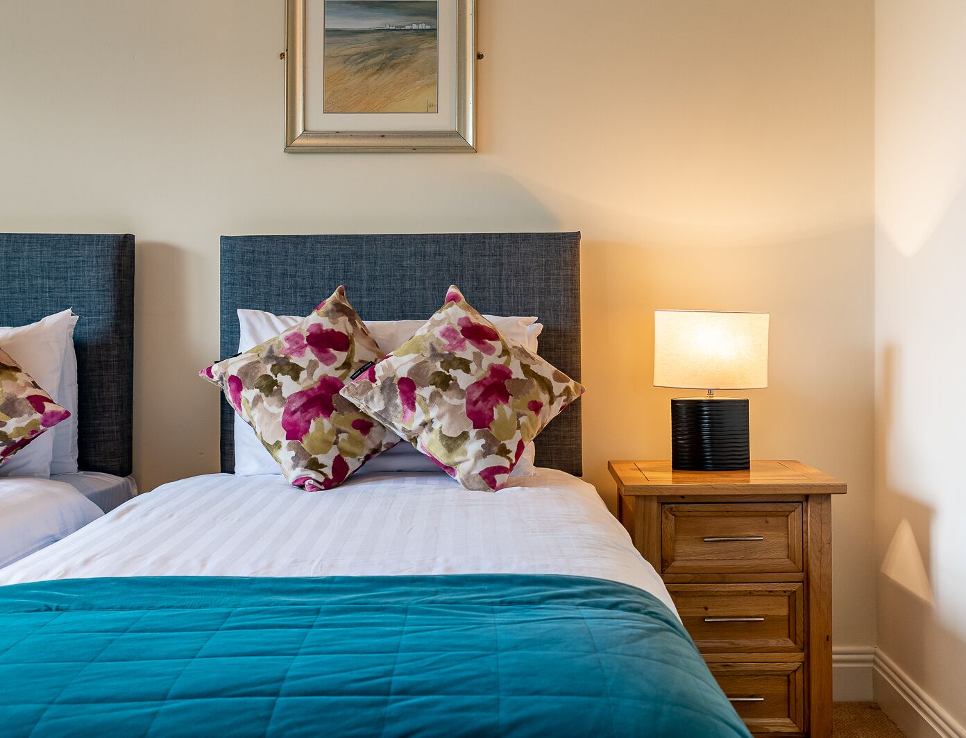 Una camera d'albergo con un letto, due cuscini decorati con un motivo floreale, un comodino con una lampada e un quadro appeso al muro.