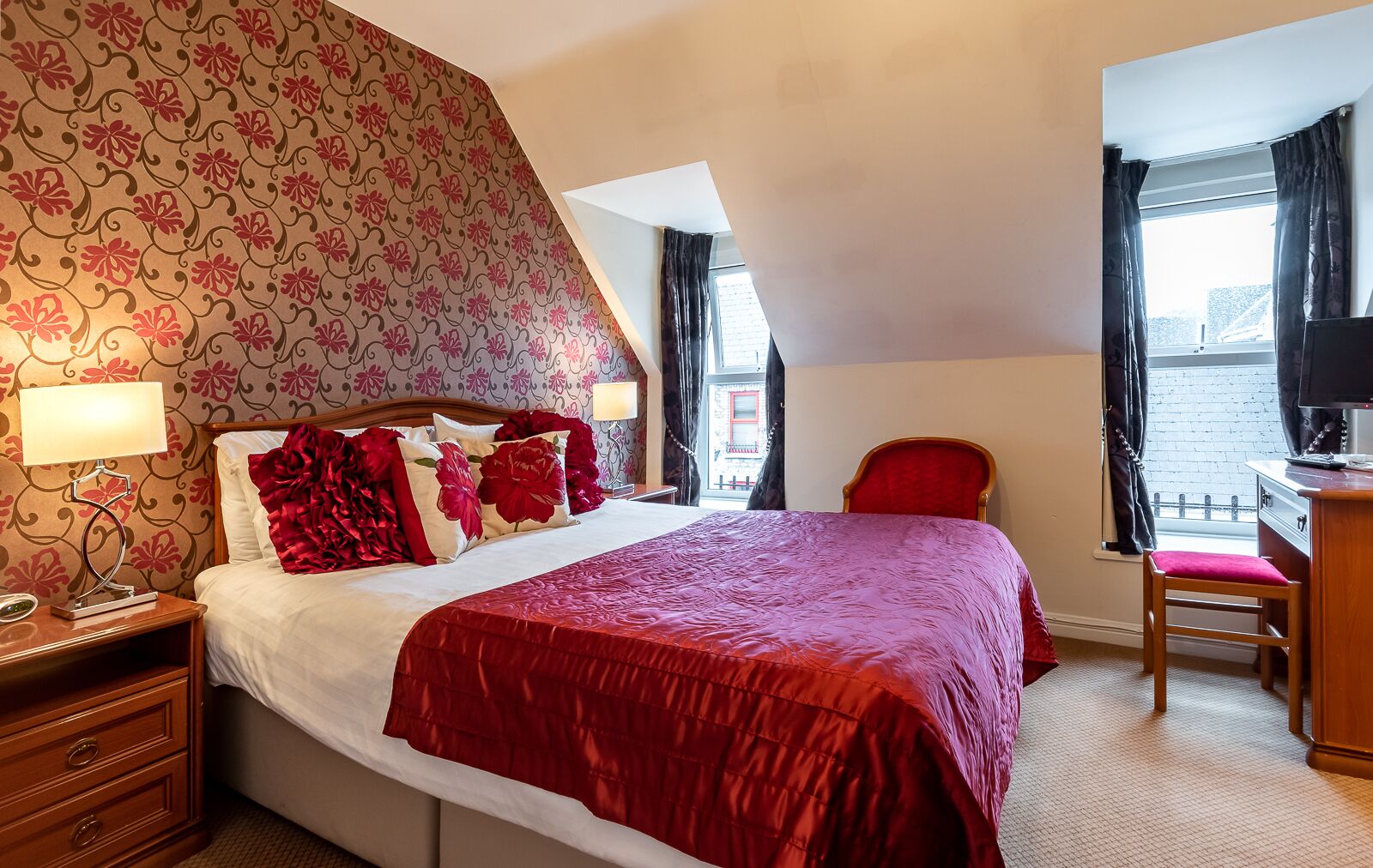 Une chambre d’hôtel avec un grand lit, un papier peint à motifs floraux, une couvre-lit rouge, une table de chevet en bois, un bureau avec une chaise et une fenêtre avec des rideaux.