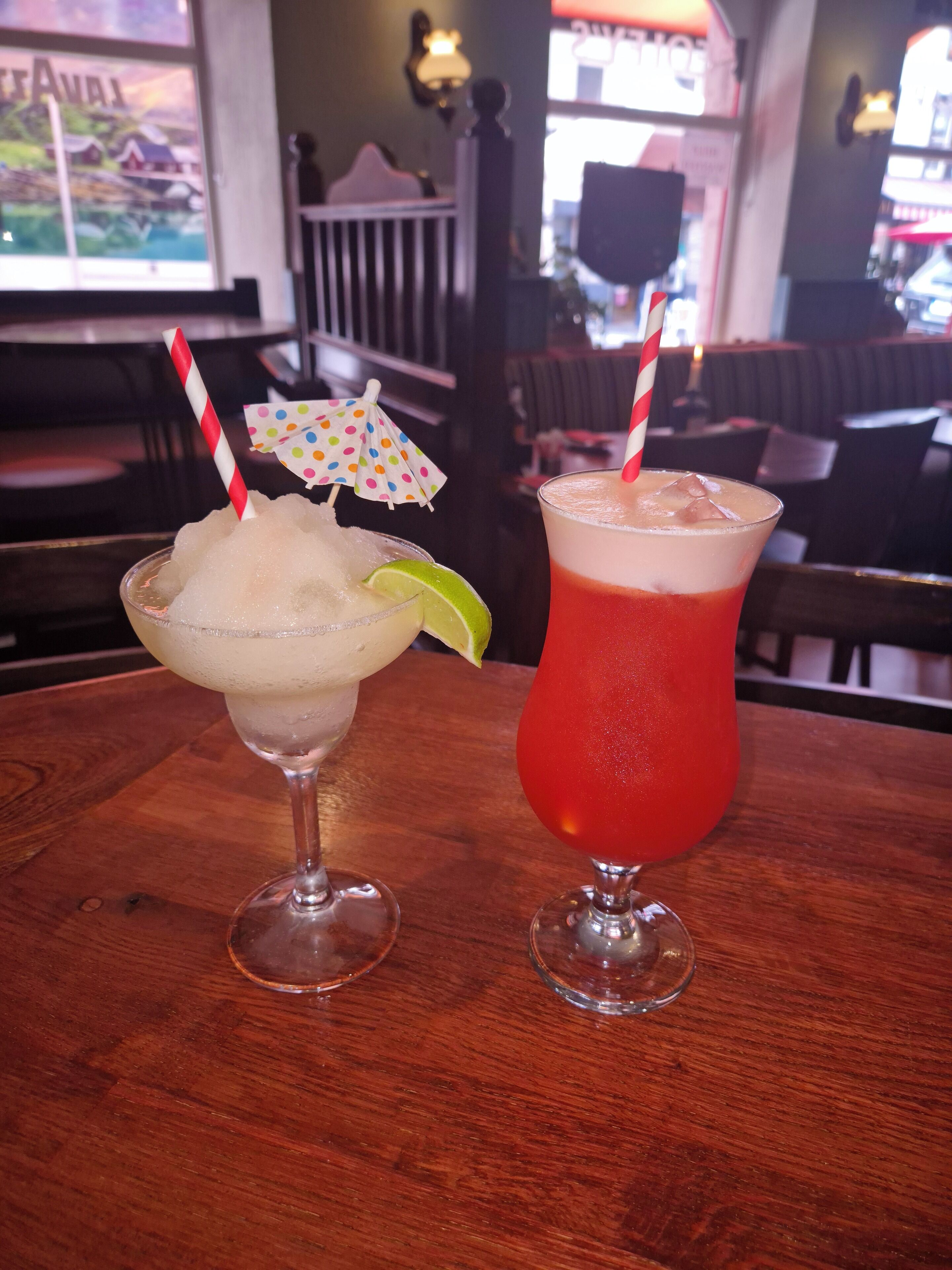 Two cocktails on a wooden table in a restaurant with a view of the outside.