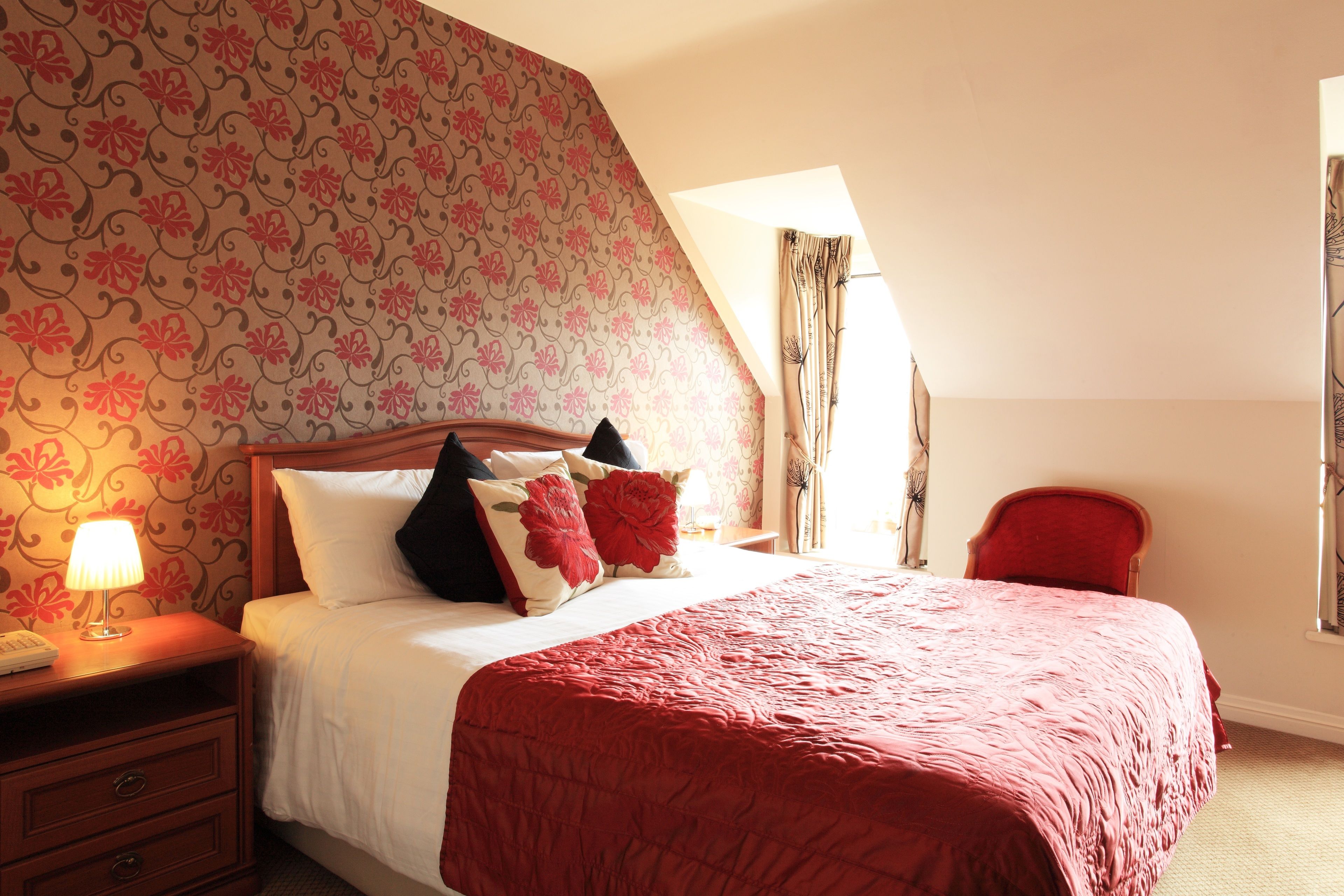 Een slaapkamer met een groot bed, bloemenbehang, een rode stoel en een nachtkastje met een lamp.