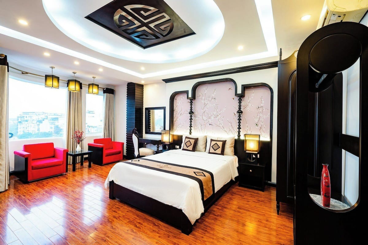 A modern hotel room with a large bed, a red armchair, a desk, and a large window offering a city view.