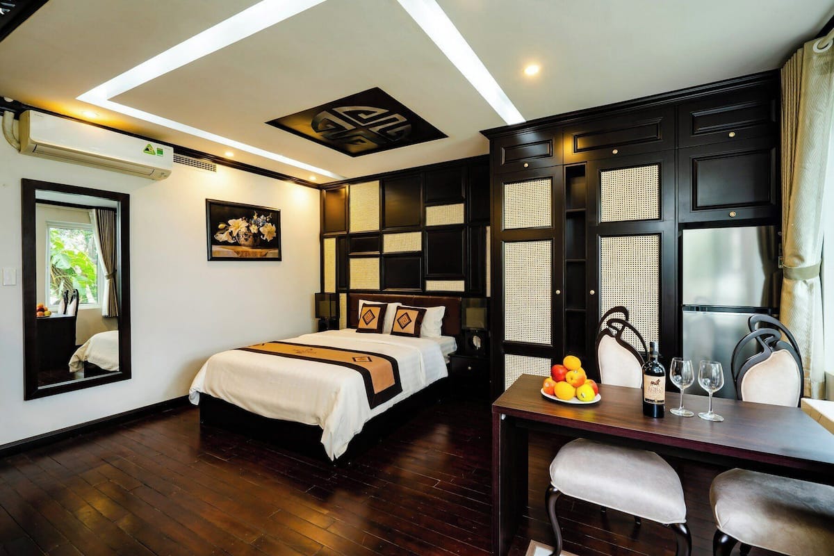 A hotel room with a bed, a desk with a fruit bowl, a wine bottle, and a mirror.