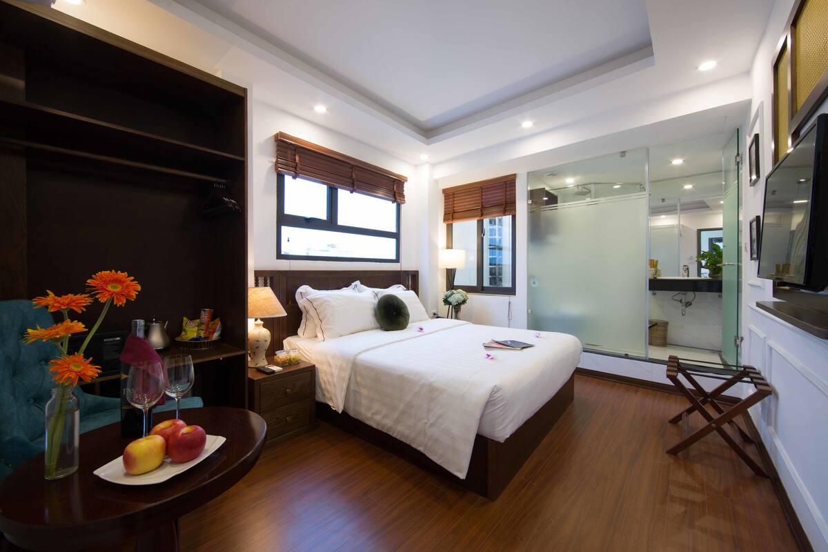A modern bedroom with a large bed, bedside tables, a nightstand, a blue armchair, a small table with fruit and flowers, and a glass-enclosed bathroom.