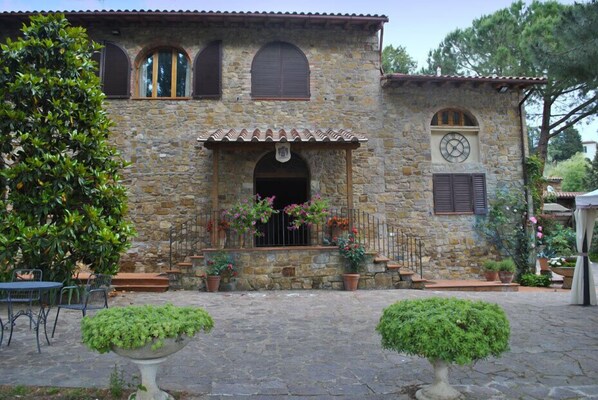 Un edificio de piedra con tejado de baldosas, un reloj y un pequeño jardín con plantas en macetas y una mesa.