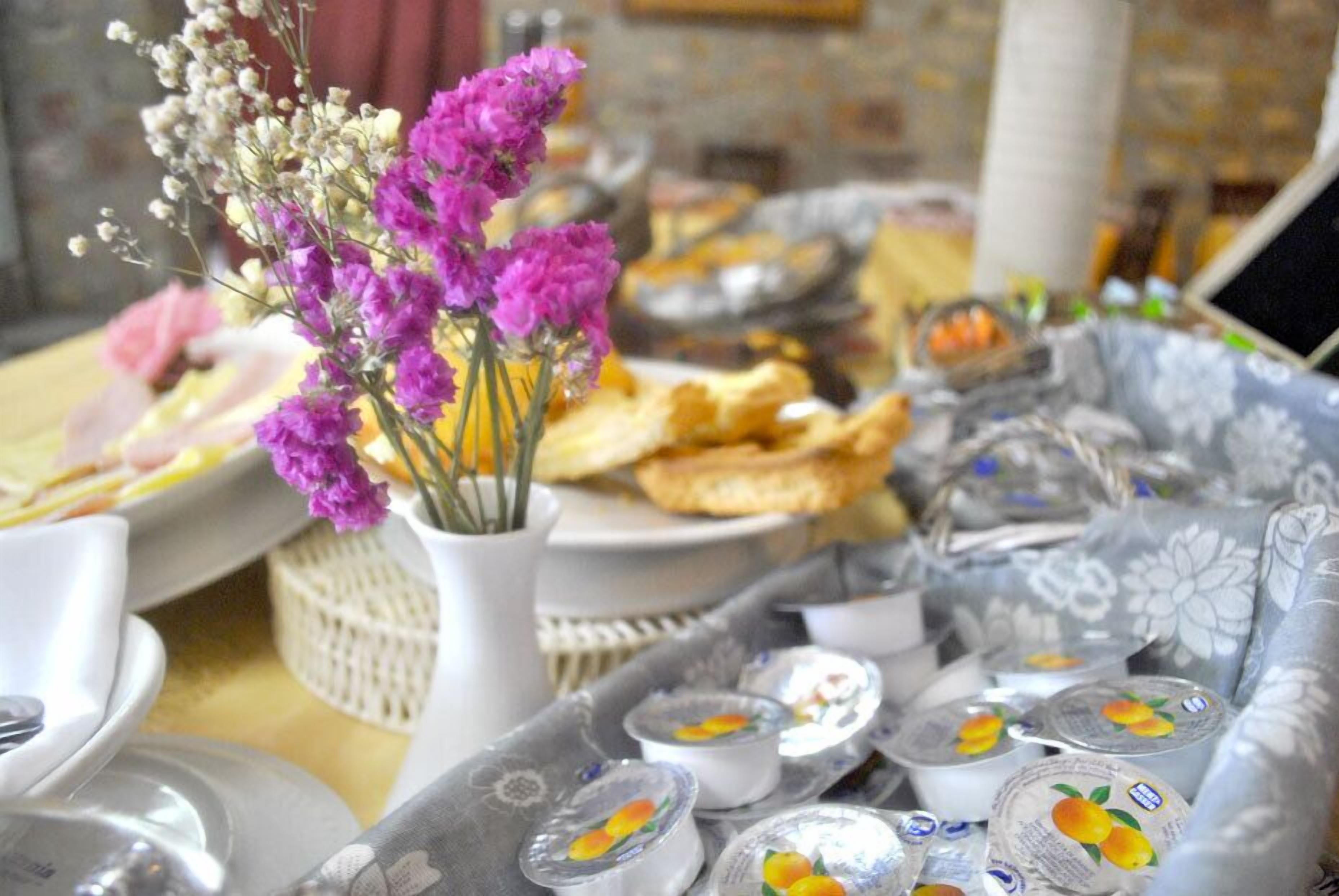 A table set with various food items, including pastries and containers of yogurt, with a vase of purple flowers in the center.