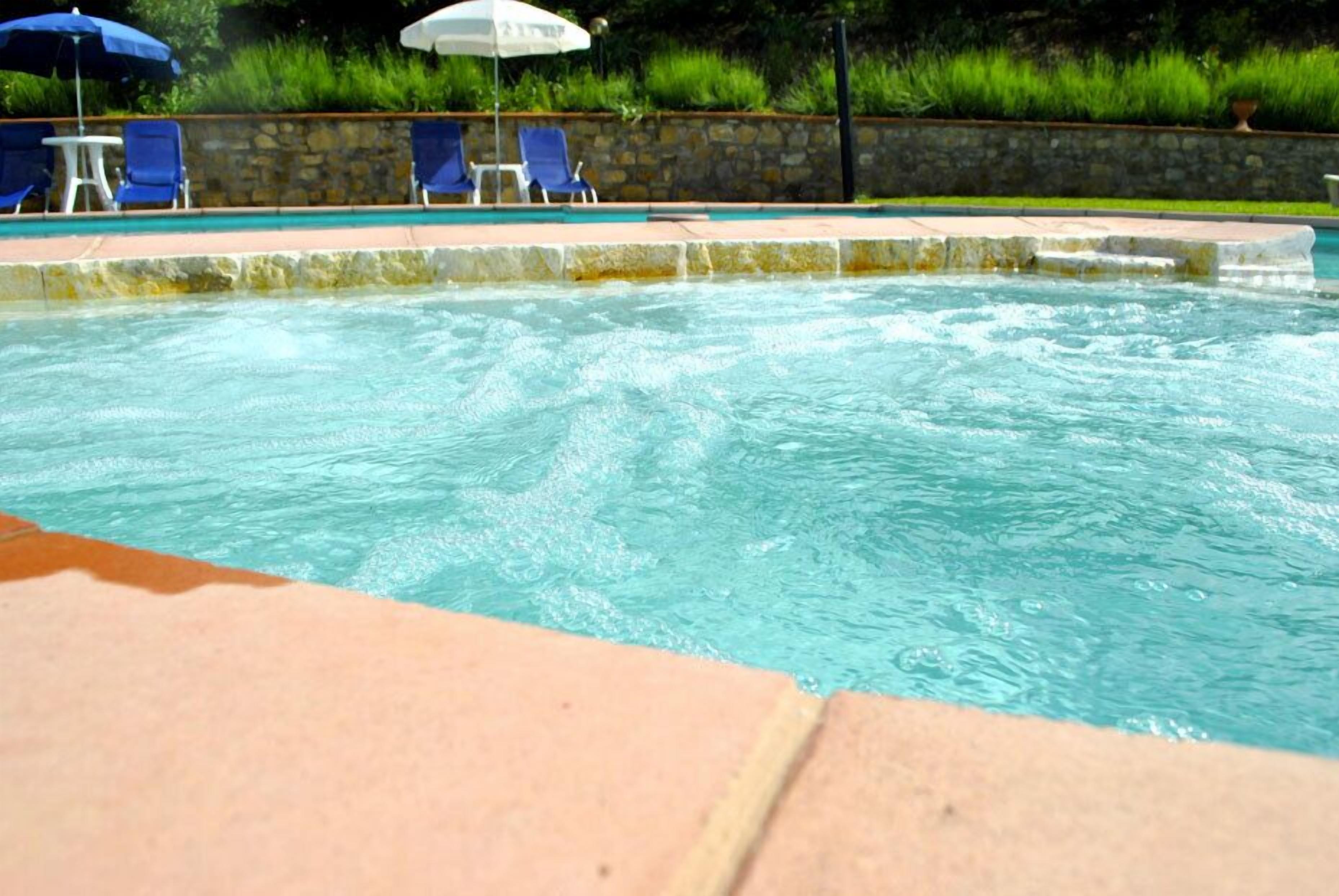 Una piscina con borde de piedra, sillas reclinables azules y sombrillas.