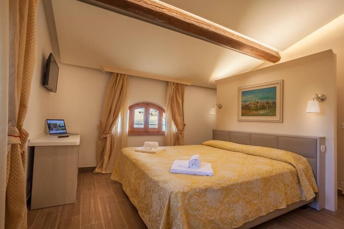 A bedroom with a bed, a desk, a laptop, a painting, and a window with curtains.