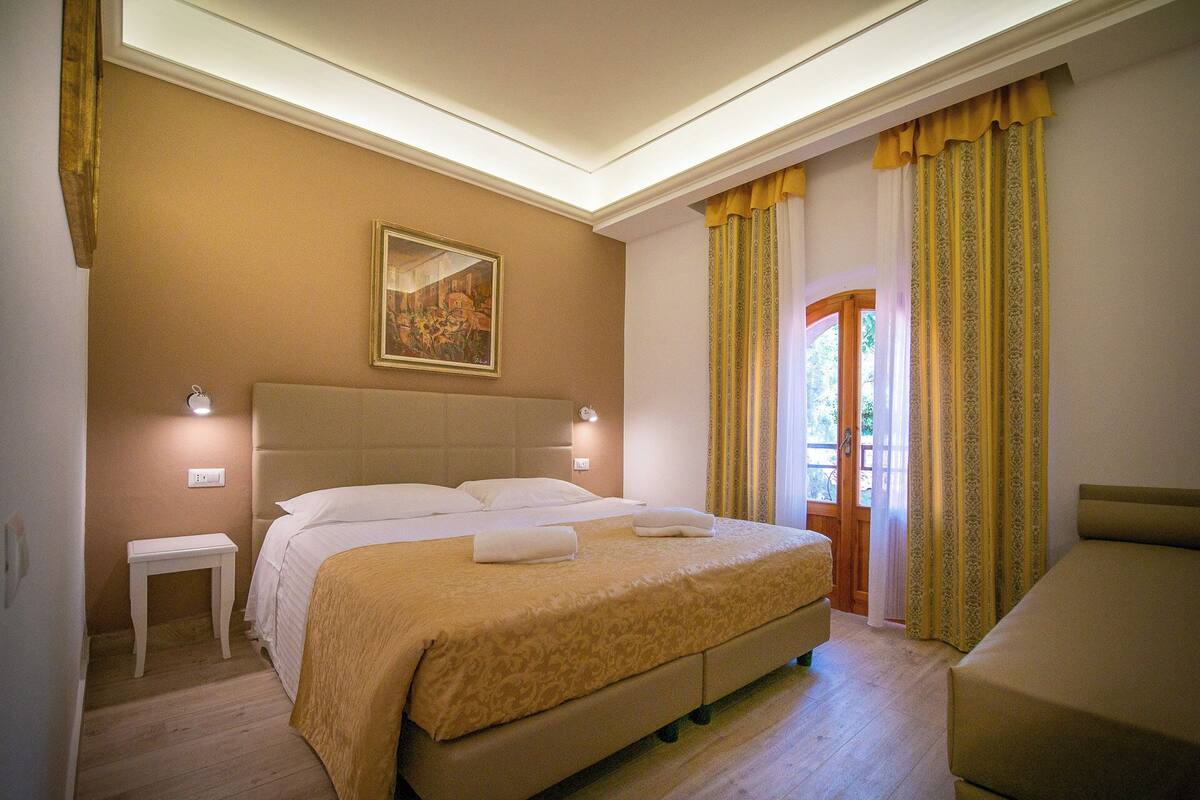 A hotel room with a large bed, a small bedside table, a painting on the wall, and a door leading to a balcony.