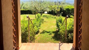 Une vue depuis un espace extérieur abrité, doté d’un sol pavé, qui donne sur un jardin où poussent différentes espèces de plantes, et sur un paysage lointain.