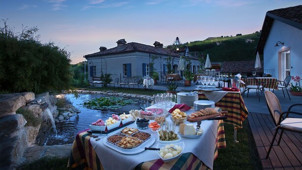 An outdoor dining area with a table set for a meal, surrounded by chairs and tables, with a building and a small stream in the background. - Agriturismo Marcarini (Neviglie)
