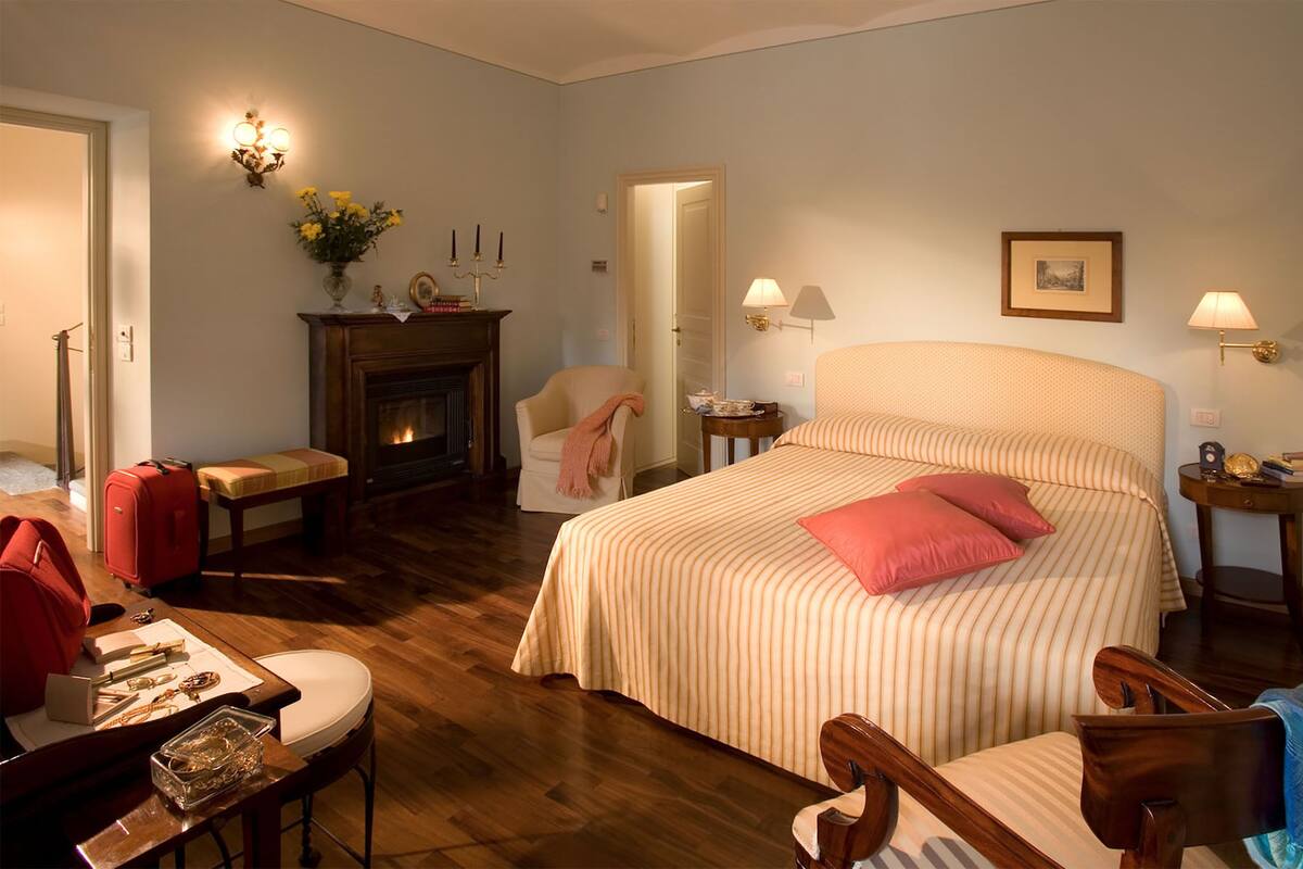 A hotel room with a large bed, a fireplace, a desk with a chair, and a small table.
