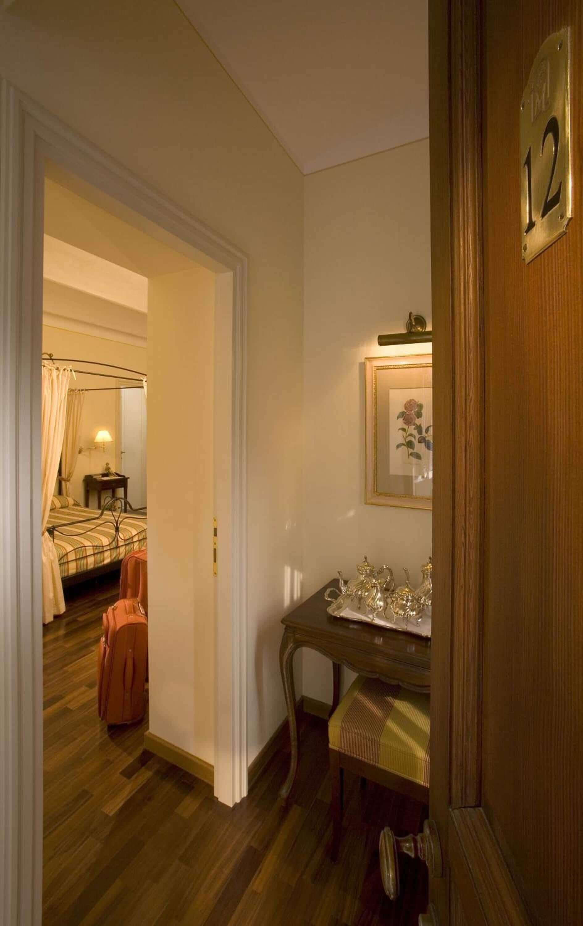 Quarto de hotel com piso de madeira, cama com roupa de cama listrada, mesa de cabeceira com abajur, espelho e cômoda com espelho em cima.