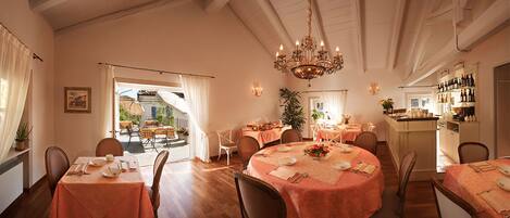 A dining area with round tables, chairs, and a chandelier. There is a view of an outdoor patio through a doorway.