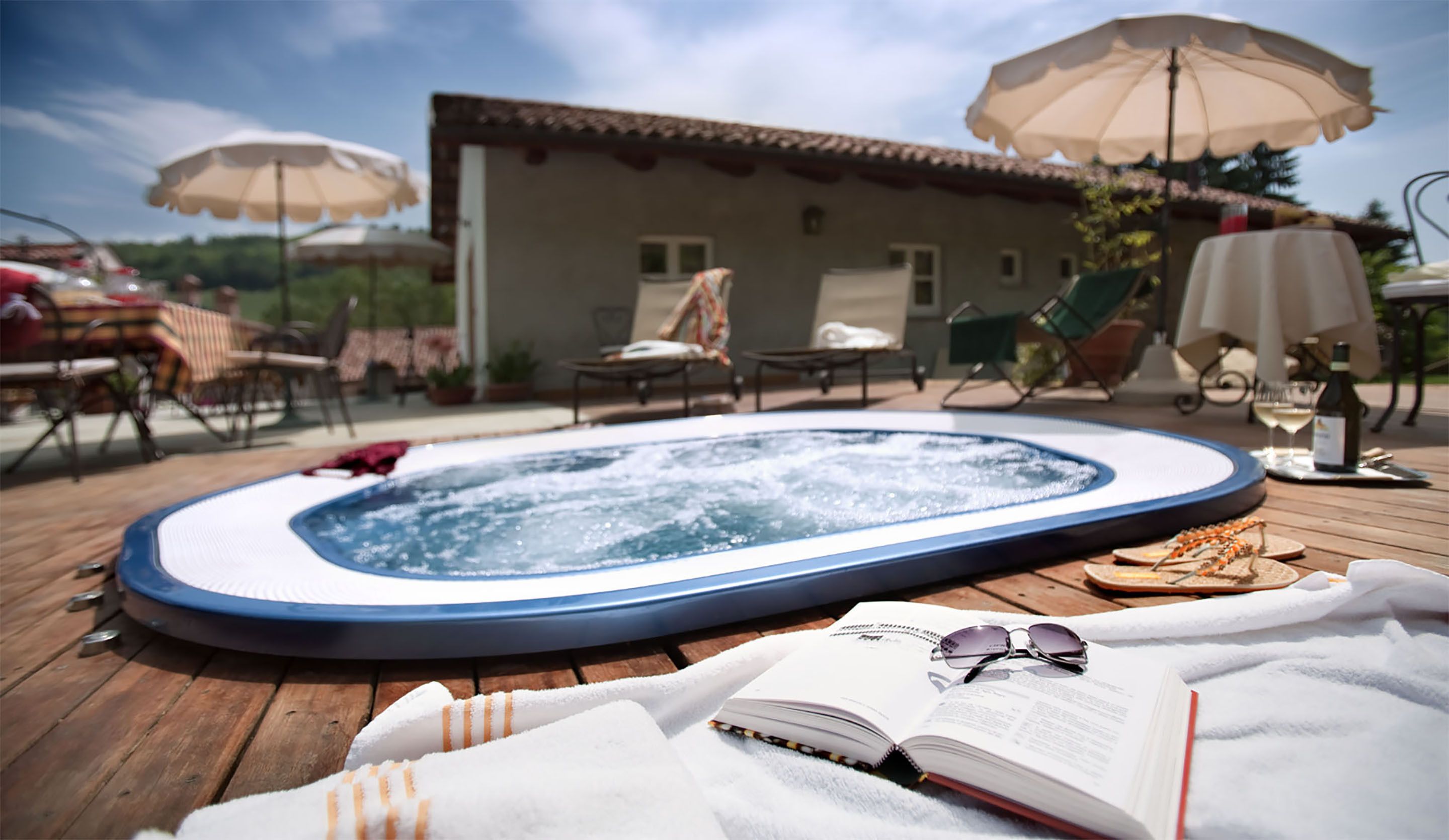 A hot tub on a wooden deck with a book and sunglasses, surrounded by outdoor furniture and umbrellas.