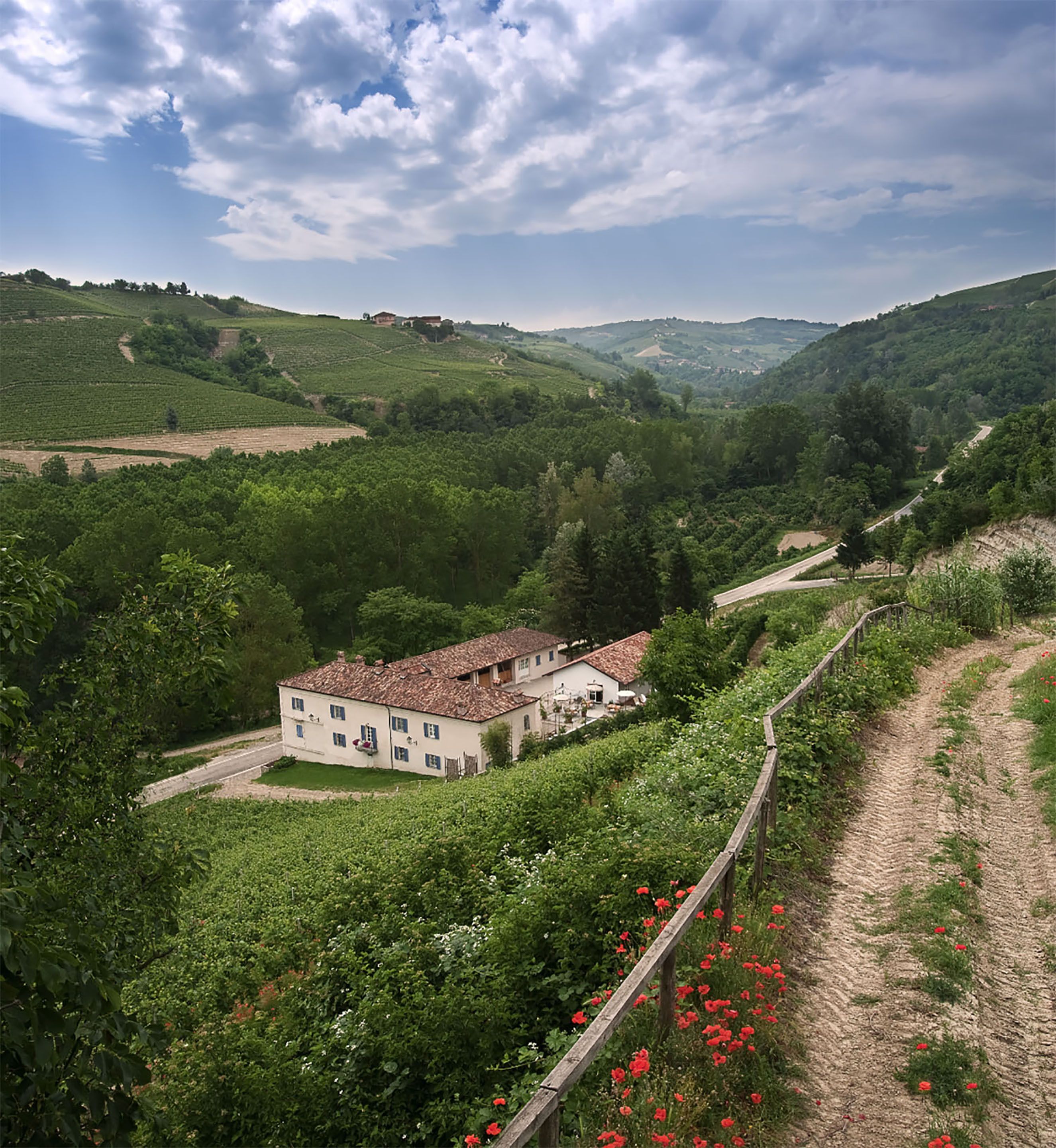 Un grand bâtiment coiffé d’un toit de tuiles rouges, entouré de vignobles verdoyants et bordé d’un chemin de terre.